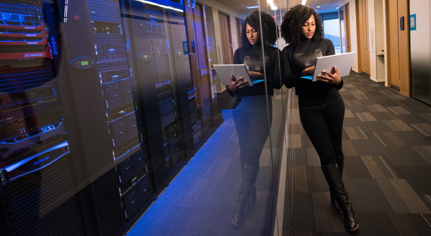 A woman stands next to a bank of servers, looking at a laptop